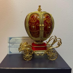 May include: A decorative Fabergé-style egg in a carriage design. The egg is red and gold with ornate detailing and a jewel-like accent. The carriage is gold-coloured with intricate scrollwork and spoked wheels. A certificate of authenticity is visible.
