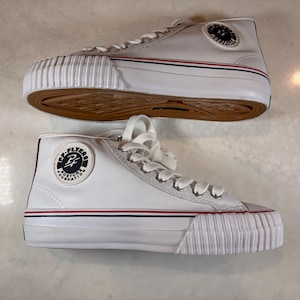Könnte beinhalten: Ein Paar weiße High-Top-Sneaker mit gerippter weißer Gummikappe, weißen Schnürsenkeln und einer gummifarbenen Sohle. An der Seite jedes Schuhs befindet sich ein kreisförmiger schwarz-weißer Aufnäher mit dem Text PF Flyers Posture Foundation.