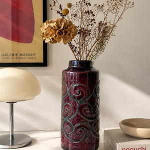 Peut inclure: Un grand vase cylindrique en céramique avec une glaçure brillante bordeaux profond, orné d'un motif répétitif de spirales en relief soulignées de bleu sarcelle. Le vase contient un bouquet de fleurs séchées, dont un hortensia et des tiges.