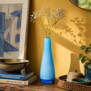 Pode incluir: Um vaso de vidro alto e esguio com um corpo liso azul-claro opaco que transita para uma base azul translúcida mais escura. O vaso tem um pescoço estreito e um fundo largo e arredondado, contendo um pequeno bouquet de flores brancas delicadas.