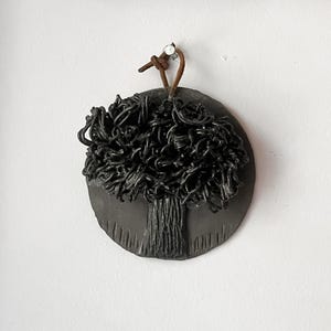 May include: A circular black wall hanging featuring a sculpted tree with a textured trunk and a dense, tangled canopy of dark wire-like branches. The piece is suspended by a brown leather cord looped through a small hole at the top edge.