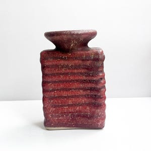 May include: A rectangular ceramic vase with a textured, deep red glaze featuring horizontal ridges across the body. The vase has a wide, flared circular opening at the top and a flat base, showcasing a rustic, hand-crafted appearance with visible variations in the red and dark brown tones throughout the surface.