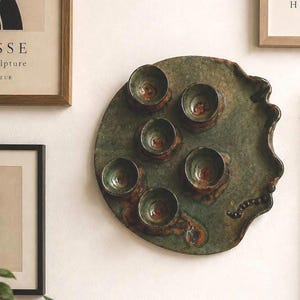 Peut inclure: Une pièce murale en céramique vert foncé de forme circulaire, comportant cinq renfoncements intégrés en forme de bol. La surface présente une texture terreuse et tachetée avec des accents bruns et un bord ondulé. Un détail perlé est fixé à droite.