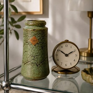 Può includere: Un vaso cilindrico in ceramica verde con una finitura craquelé testurizzata e un'etichetta a rombo Scheurich Keramik, posizionato accanto a un orologio analogico rotondo color oro con quadrante crema, lancette nere e numeri da uno a dodici.