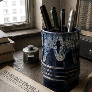 Peut inclure: Un porte-stylo cylindrique en céramique avec un vernis bleu profond et un motif de gouttes blanc sur le bord. La base présente trois nervures horizontales. Quatre stylos sont insérés, dont des instruments d'écriture noirs, verts et argentés.