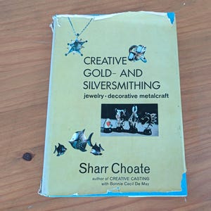 May include: A softcover book titled Creative Gold- and Silversmithing: jewelry-decorative metalcraft by Sharr Choate with Bonnie Cecil De May. The light yellow cover features illustrations of a pendant, a ring, fish, and four metal sculptures.
