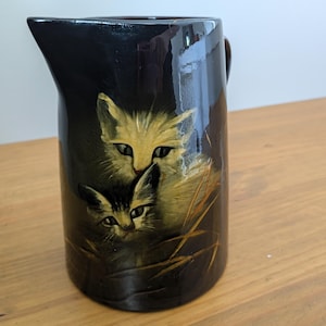 Puede incluir: Una jarra de cerámica negra brillante con una ilustración pintada de dos gatos entre hierba alta. El gato más grande es de color tostado claro con ojos oscuros, mientras que el más pequeño es oscuro con rayas. La jarra tiene asa y pico vertedor.