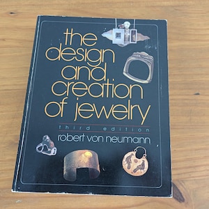 May include: A black book titled The Design and Creation of Jewelry, Third Edition by Robert von Neumann. The cover features gold text and illustrations of various jewelry pieces, including a ring, a cuff bracelet, and a circular pendant.