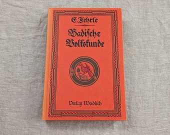 Pode incluir: Um livro de capa dura com uma capa laranja vibrante, apresentando texto preto e um emblema circular. O título diz Badische Volkskunde de E. Fehrle, com Verlag Weidlich na parte inferior. Uma borda geométrica preta emoldura a capa.