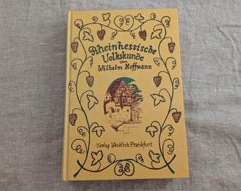 Pode incluir: Um livro de capa dura com capa bege apresentando uma borda de videira verde-escura com cachos de uvas vermelhos. O centro exibe um desenho de um edifício com o texto Rheinhessische Volkskunde von Wilhelm Hoffmann e Verlag Weidlich Frankfurt.