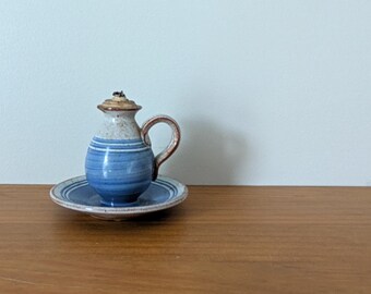 May include: A small ceramic oil lamp with a handle, featuring a speckled cream-colored top and a blue-striped base. The lamp sits on a matching circular saucer with blue concentric rings. The design includes a small wick protruding from the top.