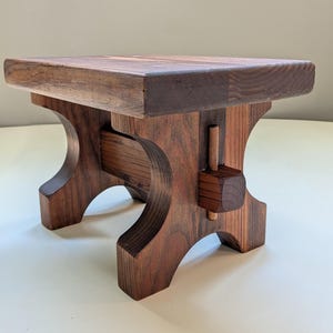 May include: A handcrafted wooden stool with a dark brown finish. The stool features a square top and curved legs, showcasing a unique design. The dimensions are approximately 12 inches in height and 14 inches in width.