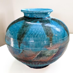 Op de afbeelding: Een grote, ronde keramische vaas met een brede opening. De vaas is voornamelijk turquoise met donkerblauwe en bruine accenten. Het oppervlak heeft een gekraakte textuur, wat visuele interesse toevoegt aan het decoratieve stuk.