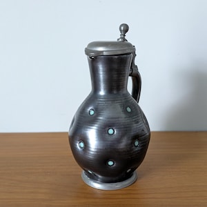 May include: A dark brown ceramic pitcher with a rounded body and a metallic lid. The pitcher features a series of small, circular indentations across its surface, each containing a light blue dot. It has a handle and a metal base rim.