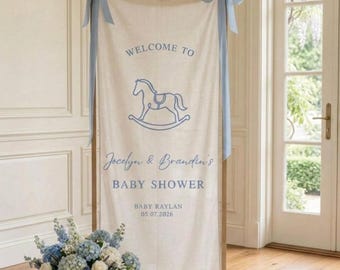 May include: A vertical cream-coloured fabric banner hanging from a wooden frame, decorated with light blue ribbons. It features a blue rocking horse graphic, the text "WELCOME TO", "Jocelyn & Brandin's BABY SHOWER", "BABY RAYLAN", and "05.07.2026".