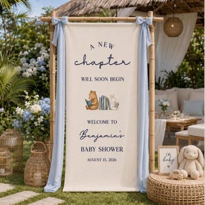 May include: A cream-colored fabric banner hanging on a bamboo frame with light blue bows. The text reads A NEW chapter WILL SOON BEGIN, WELCOME TO Benjamin's BABY SHOWER, AUGUST 15, 2026, with illustrations of a bear and rabbit reading books.