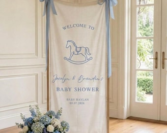 May include: A vertical cream-colored fabric banner suspended from a wooden frame with light blue ribbons. The banner features a blue rocking horse graphic and text reading WELCOME TO, JOCELYN & BRANDIN'S BABY SHOWER, BABY RAYLAN, 05.07.2026.