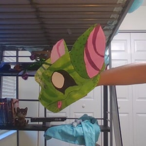 May include: A green creature mask featuring large pointed ears with pink inner details, dark eye patches, and a small pink tongue. The mask is constructed from a textured material and is designed to be worn as a hand puppet or headpiece.