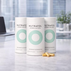 May include: Three white cylindrical bottles of Nutrafol Women's Balance hair growth supplement. Each bottle has a teal circular design and text. Several gold capsules are in front of the bottles. The background is blurred.