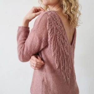 May include: A dusty rose sweater with a deep V-back and long sleeves. The back features a decorative, textured design resembling a feather, embellished with small, scattered beads. The sweater is made of a soft, knitted material.
