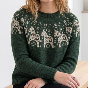 Könnte beinhalten: Ein dunkelgrüner Pullover mit Rundhalsausschnitt und langen Ärmeln. Der Pullover zeigt ein Muster aus beigefarbenen Dala-Pferden und schneeflockenartigen Motiven auf der Brust und den oberen Ärmeln. Das Strickwerk hat gerippte Bündchen und einen Saum.