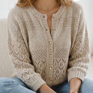 Op de afbeelding: Beige gebreid vest met een ronde halslijn en gouden knopen. De mouwen hebben een bladachtig ajourmotief. Het vest heeft een lichte, neutrale kleur en een klassieke stijl met knopen.