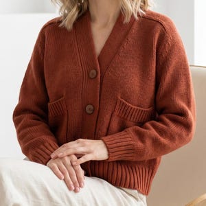 May include: A rust-coloured cardigan sweater with a V-neck, two large pockets, and two brown buttons. The sweater has a ribbed hem and cuffs. The model is wearing cream-coloured trousers.