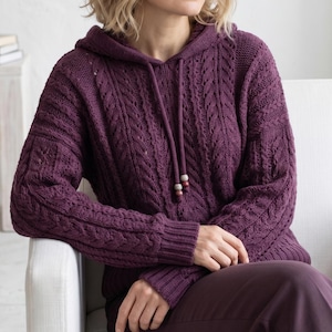 Op de afbeelding: Een pruimkleurige hoodie met een kabelgebreid ontwerp. De trui heeft een trekkoord met houten kralen en geribde manchetten. Het model draagt de trui terwijl ze op een witte stoel zit.