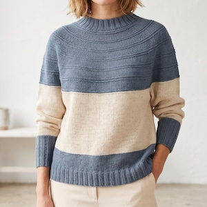 May include: A blue and beige color-block sweater with a ribbed neckline, cuffs, and hem. The top portion of the sweater is blue with horizontal ribbed detailing, while the midsection is beige. The sleeves and lower hem are blue.