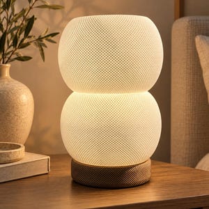 May include: A table lamp featuring two stacked, spherical white glass shades with a fine, textured grid pattern. The lamp is supported by a circular base wrapped in a tan, woven fabric material that emits a warm, soft glow when illuminated.