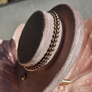 May include: A bracelet composed of a single strand of polished, metallic rose gold-toned spherical beads. The beads are uniform in size and arranged in a continuous circular formation, reflecting light across their smooth, rounded surfaces.