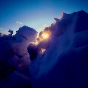 Op de afbeelding: Een dramatische landschapsfoto met besneeuwde toppen onder een heldere, diepblauwe lucht. De zon schijnt fel door een opening in de sneeuw en creëert een stralend effect. De afbeelding wordt gedomineerd door koele blauw- en wittinten.