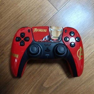 Peut inclure: Une manette de jeu vidéo rouge et noire arborant un graphisme d'Iron Man, le mot Avengers et un logo Avengers. La manette comprend des joysticks analogiques noirs, une croix directionnelle et des boutons d'action avec des symboles triangle, cercle, croix et carré.