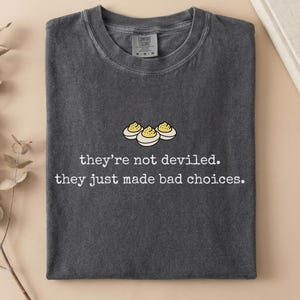 May include: A charcoal gray crew neck t-shirt featuring a graphic of three halved deviled eggs and the white text: they're not deviled. they just made bad choices. The shirt has a visible brand label at the inner neckline and a short-sleeve design.