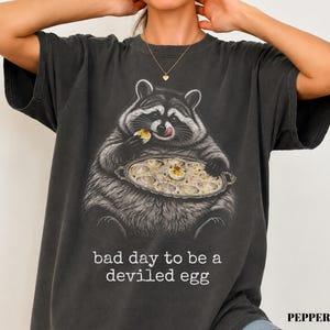 May include: A charcoal gray t-shirt featuring a graphic illustration of a raccoon eating a deviled egg from a platter. Below the raccoon, the text reads, "bad day to be a deviled egg." The shirt has short sleeves and a crew neckline, with a vintage-style wash.