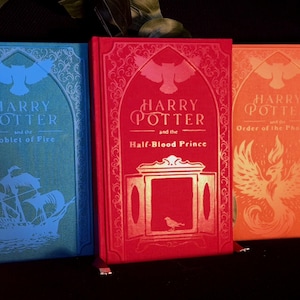 May include: Three hardback books titled Harry Potter and the Goblet of Fire in blue, Harry Potter and the Half-Blood Prince in red, and Harry Potter and the Order of the Phoenix in orange, each featuring gold foil illustrations and text.