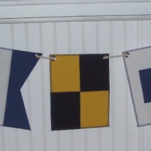 May include: A nautical flag banner with five colorful signal flags. The flags are in various shapes and feature blue, white, red, and yellow designs. The flags are strung together on a white rope.