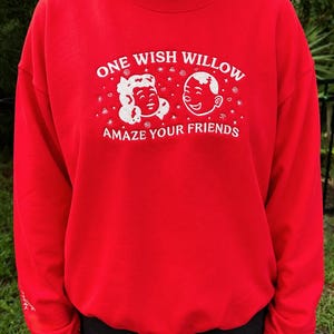 Puede incluir: Una sudadera roja de cuello redondo con texto bordado en blanco que dice ONE WISH WILLOW y AMAZE YOUR FRIENDS. Entre las l&iacute;neas de texto hay dos caras de dibujos animados bordadas en blanco rodeadas de peque&ntilde;as estrellas y espirales.