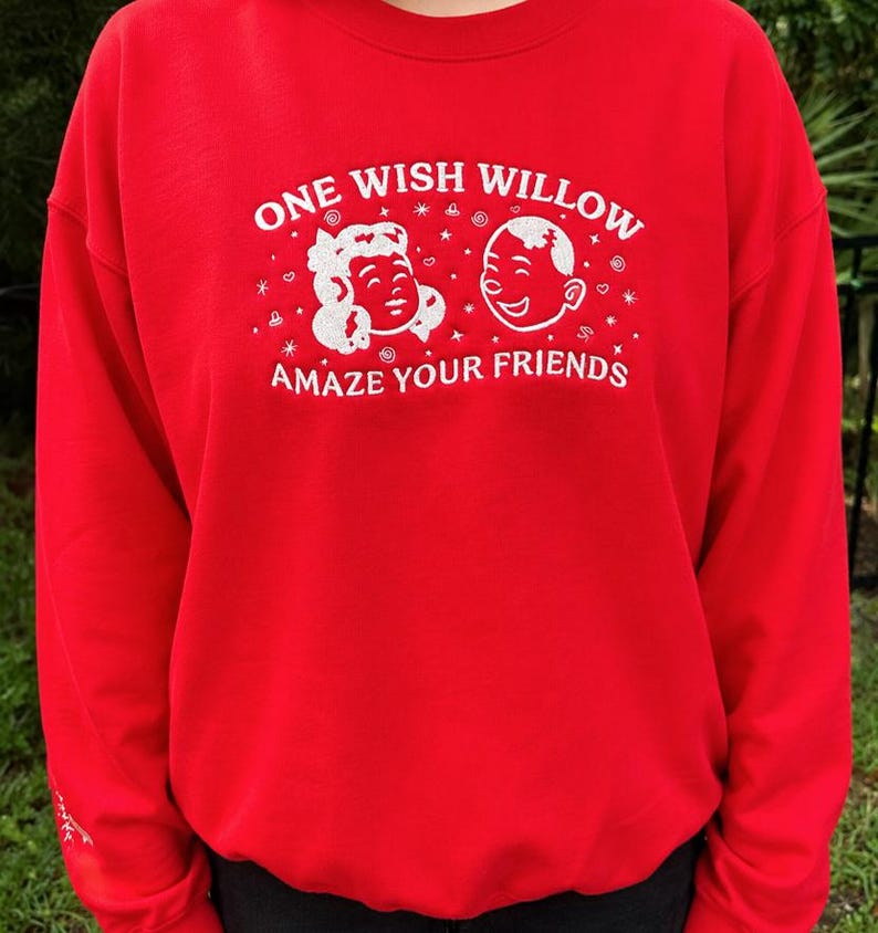 Puede incluir: Una sudadera roja de cuello redondo con texto bordado en blanco que dice ONE WISH WILLOW y AMAZE YOUR FRIENDS. Entre las l&iacute;neas de texto hay dos caras de dibujos animados bordadas en blanco rodeadas de peque&ntilde;as estrellas y espirales.