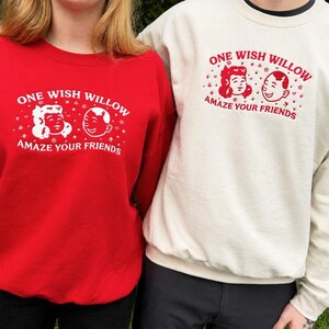 Puede incluir: Una sudadera roja y una sudadera color crema, ambas con un estampado gr&aacute;fico de dos caras de dibujos animados y el texto "ONE WISH WILLOW AMAZE YOUR FRIENDS". La sudadera roja tiene texto blanco y la sudadera color crema tiene texto rojo.