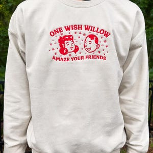 Puede incluir: Una sudadera de cuello redondo color crema con bordado rojo en el pecho. El dise&ntilde;o incluye el texto ONE WISH WILLOW y AMAZE YOUR FRIENDS, rodeando un gr&aacute;fico de dos caras sonrientes con peque&ntilde;os detalles de estrellas y espirales.
