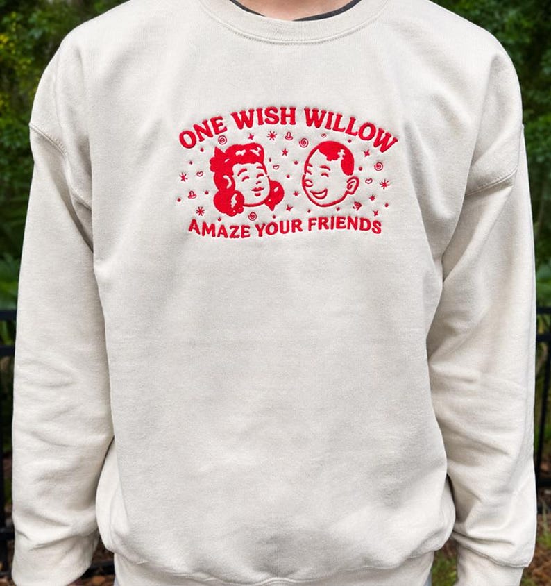 Puede incluir: Una sudadera de cuello redondo color crema con bordado rojo en el pecho. El dise&ntilde;o incluye el texto ONE WISH WILLOW y AMAZE YOUR FRIENDS, rodeando un gr&aacute;fico de dos caras sonrientes con peque&ntilde;os detalles de estrellas y espirales.