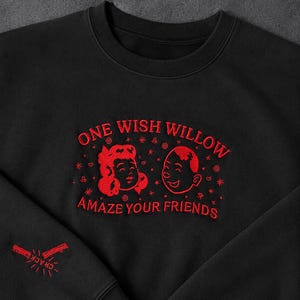 Puede incluir: Una sudadera negra de cuello redondo con bordado rojo en el pecho que dice ONE WISH WILLOW AMAZE YOUR FRIENDS, acompa&ntilde;ada de dos caras de dibujos animados y peque&ntilde;os iconos de estrellas. La manga presenta un gr&aacute;fico bordado en rojo con la palabra CRACK.
