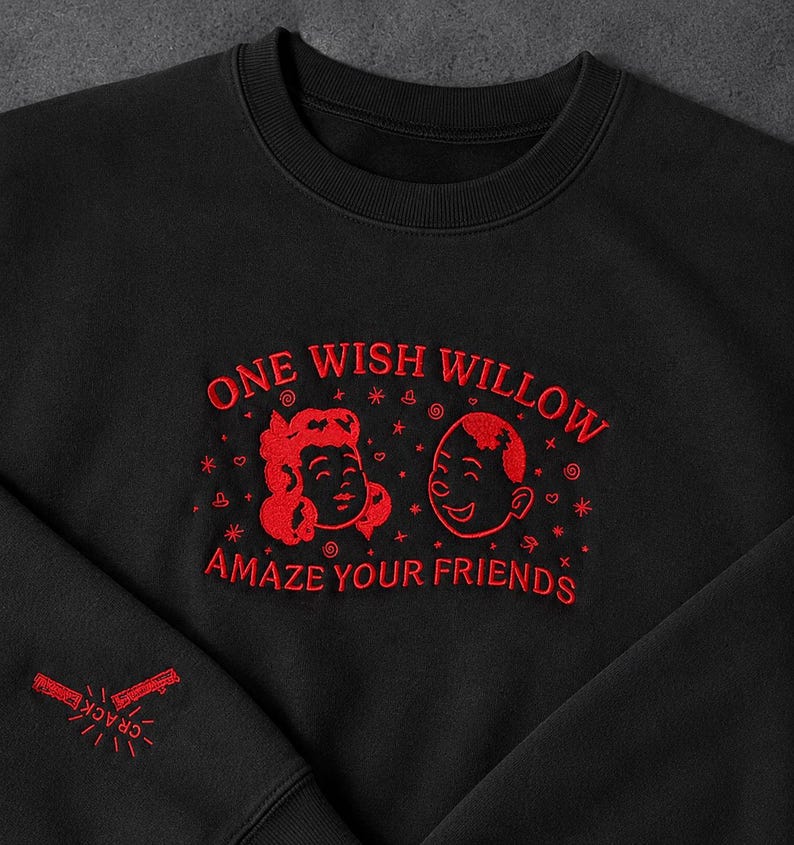 Puede incluir: Una sudadera negra de cuello redondo con bordado rojo en el pecho que dice ONE WISH WILLOW AMAZE YOUR FRIENDS, acompa&ntilde;ada de dos caras de dibujos animados y peque&ntilde;os iconos de estrellas. La manga presenta un gr&aacute;fico bordado en rojo con la palabra CRACK.