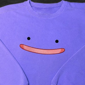 May include: A light purple crewneck sweatshirt featuring an embroidered graphic on the chest. The design consists of two small black circular eyes positioned above a wide, curved, light pink mouth with a thin dark red outline.