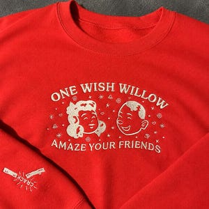 Puede incluir: Una sudadera de cuello redondo en color rojo brillante con bordados en blanco. El pecho muestra el texto ONE WISH WILLOW y AMAZE YOUR FRIENDS rodeando dos caras de dibujos animados. La manga presenta un gr&aacute;fico bordado de un hueso de la suerte con la palabra CRACK.