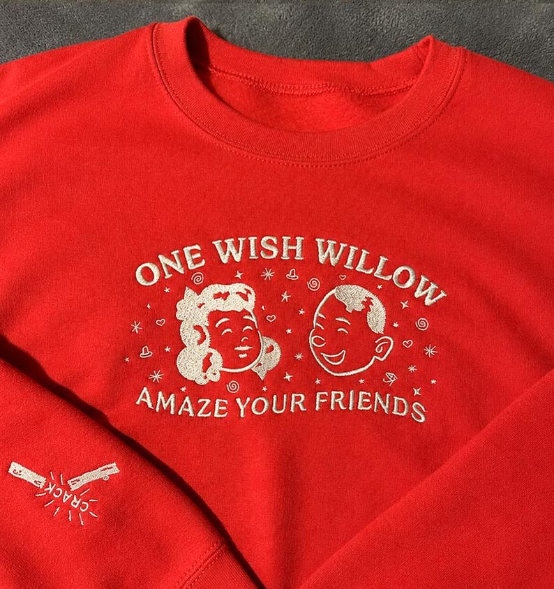 Puede incluir: Una sudadera de cuello redondo en color rojo brillante con bordados en blanco. El pecho muestra el texto ONE WISH WILLOW y AMAZE YOUR FRIENDS rodeando dos caras de dibujos animados. La manga presenta un gr&aacute;fico bordado de un hueso de la suerte con la palabra CRACK.