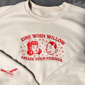 May include: Cream-colored sweatshirt with red embroidered text and graphics. The design includes the words "ONE WISH WILLOW" above two cartoon faces and the phrase "AMAZE YOUR FRIENDS" below. A small embroidered graphic of a firecracker is on the sleeve.