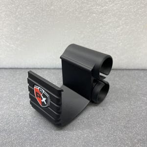 May include: A black, 3D-printed bracket with a shield logo featuring red and white accents. The bracket has a curved top and two circular openings. The base of the bracket is flat and has a ridged texture.