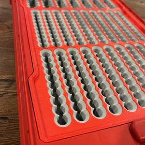 Puede incluir: Una caja de aparejos de pesca con mosca de plástico rojo con un interior de espuma gris. El interior tiene múltiples filas de ranuras y ranuras para guardar moscas de pesca. La caja está abierta, revelando el interior.